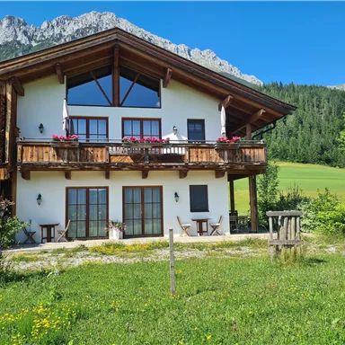 Ein gemütliches Holzhaus mit einem großen Balkon und Blumenkästen. Im Hintergrund sind grüne Wiesen und Berge zu sehen.
