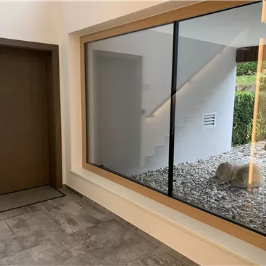 Ein moderner Eingangsbereich mit einer großen Glasscheibe und einem Blick auf einen kleinen Garten. Die Wände sind hell und die Tür ist aus Holz.