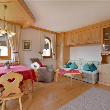 Ein gemütliches Wohnzimmer mit einem großen Tisch, Stühlen und einer hellen Einrichtung aus Holz. Es gibt eine Sofaecke mit Kissen und großen Fenstern, die viel Licht hereinlassen.