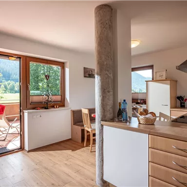 A bright and inviting kitchen with wooden cabinets and modern appliances. In the background, there are windows overlooking the countryside and a table on the terrace.