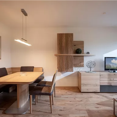 A modern dining room with a large table and comfortable chairs. There is a television on the wall and a decorative element.
