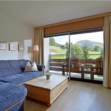 A bright living room with a blue sofa and a wooden table. Large windows offer a view of the mountains and the green landscape outside.