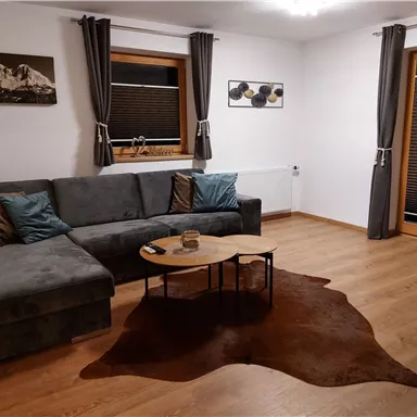 A cozy living room with a gray sofa and a round table. Large windows provide a bright space, and the wooden floors give a warm atmosphere.