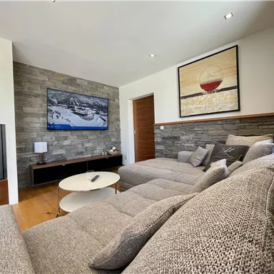 Ein modernes Wohnzimmer mit einer großen grauen Couch und einem Fernseher an der Wand. Es gibt einen runden Tisch und dekorative Elemente wie ein Gemälde.