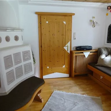 Ein gemütlicher Wohnraum mit Holzdekor und einer angrenzenden Tür. Auf der Sitzbank liegen Kissen und ein Teppich auf dem Boden.