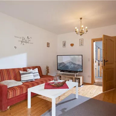 Ein gemütliches Wohnzimmer mit einem roten Sofa und einem weißen Tisch. An der Wand hängt ein Fernseher, und es gibt Pflanzen sowie eine warm beleuchtete Atmosphäre.