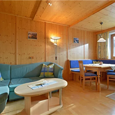 Ein gemütlicher Wohnraum mit Holzverkleidung, einem blauen Sofa und einem Esstisch. Große Fenster sorgen für viel Tageslicht.