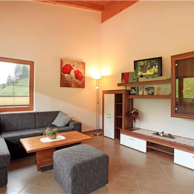 Ein modernes Wohnzimmer mit einer grauen Couch, einem Holzkaffee-Tisch und einem passenden Hocker. An der Wand sind Fenster und einige dekorative Elemente zu sehen.