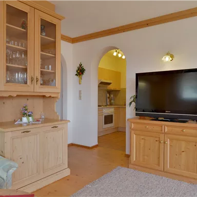 Ein gemütliches Wohnzimmer mit hellem Holz und einem Fernseher. Im Hintergrund ist die offene Küche sichtbar.