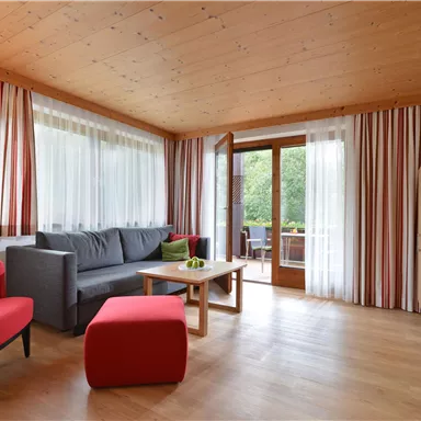 Ein gemütliches Wohnzimmer mit einem grauen Sofa und roten Stühlen. Große Fenster sorgen für viel Tageslicht und einen Blick auf den Balkon.