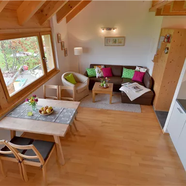 Ein modernes Wohnzimmer mit großen Fenstern und Holzbalkendecke. Der Raum ist gemütlich eingerichtet mit einer Couch, einem Tisch und einer offenen Küche.