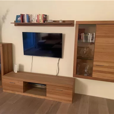Ein moderner Wohnraum mit einem Fernseher an der Wand und einem Holzregal darunter. Neben dem Fernseher befindet sich ein eleganter Schrank mit Glastüren.