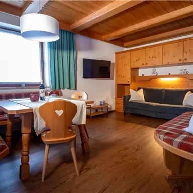 A cozy living room with a wooden ceiling and warm colors. There is a dining table with chairs, a cozy sitting area, and a TV wall.