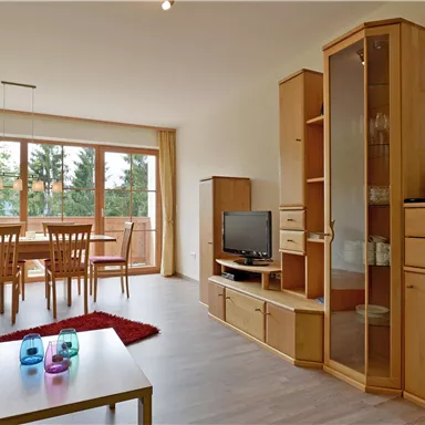 Ein helles Wohnzimmer mit Holzmöbeln und großen Fenstern. In der Mitte steht ein Tisch mit bunten Gläsern und einem roten Teppich.