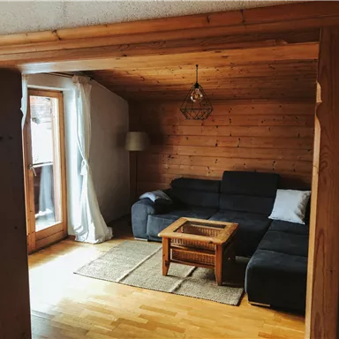 Ein gemütliches Wohnzimmer mit einer dunklen Couch und einem Holztisch. Das Zimmer hat große Fenster, die viel Licht hereinlassen.