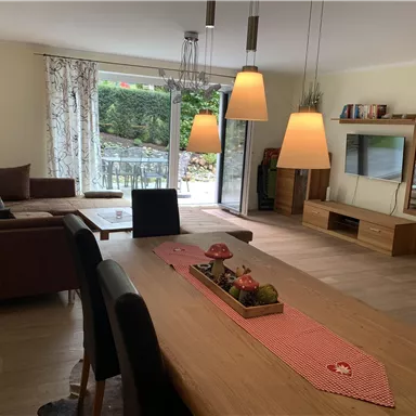 Ein modernes Wohnzimmer mit einer gemütlichen Couch und einem Esstisch. Große Fenster bringen viel Licht und bieten einen Blick auf den Garten.