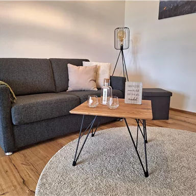 Ein gemütliches Wohnzimmer mit einem grauen Sofa und einem Holztisch. Auf dem Tisch stehen Gläser und eine Dekoration.
