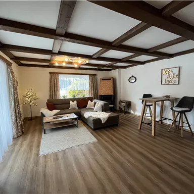 Ein gemütliches Wohnzimmer mit Holzbalken und modernen Möbeln. Große Fenster lassen viel Licht herein und sorgen für eine einladende Atmosphäre.