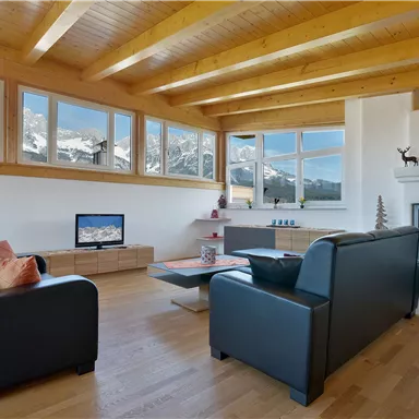 Ein modernes Wohnzimmer mit zwei schwarzen Sofas und Holzdecke. Große Fenster bieten einen Blick auf bergige Landschaften.