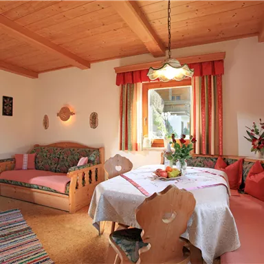 Ein gemütlicher Wohnraum mit Holzdecke und bunten Möbeln. Ein runder Tisch ist mit Blumen dekoriert, und große Fenster lassen viel Licht herein.