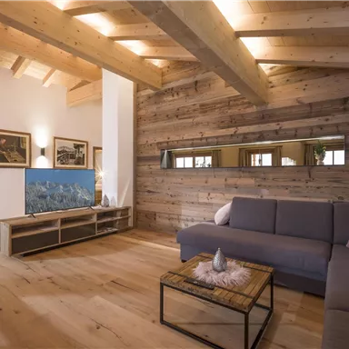 A modern living room with a wooden ceiling and floor. A comfortable couch is positioned in front of a TV cabinet that displays a television.