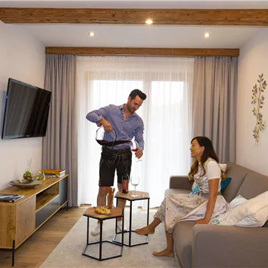 A cozy living space with a man pouring wine and a woman relaxing on the couch. The room is modern and bright with wooden details.