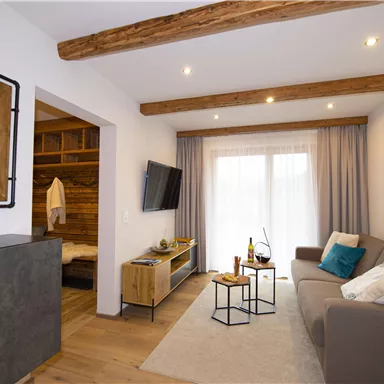 A cozy living room with wooden ceiling beams and modern furnishings. A sofa, a coffee table, and large windows create a bright, inviting atmosphere.