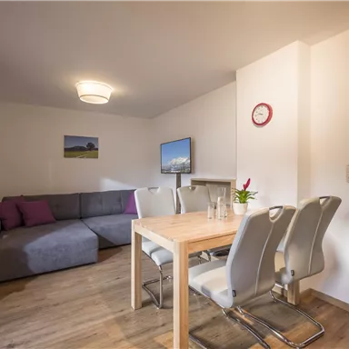 A bright living room with a gray couch and a wooden table. The room has a modern style and large windows with natural light.