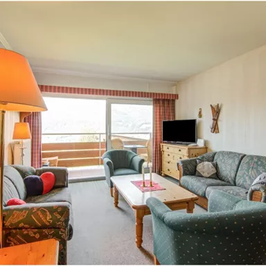 A cozy living room with comfortable sofas and a wooden table. Large windows offer a view of the terrace and the landscape.