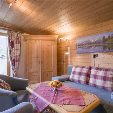 A cozy room with wooden walls and a large window. On the table is a fruit bowl, and a blue sofa with checkered cushions invites you to relax.