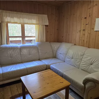 Eine gemütliche Sitzecke mit einer hellen Ledercouch und einem Holztisch. Der Raum ist mit Holzvertäfelung und einem Fenster mit Vorhängen gestaltet.