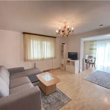 Ein modernes Wohnzimmer mit einem grauen Sofa, einem Couchtisch und einem Fernseher. Große Fenster sorgen für viel Tageslicht und eine helle Atmosphäre.