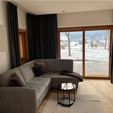Ein gemütliches Wohnzimmer mit einem grauen Sofa und modernen Möbeln. Große Fenster bieten einen Blick auf eine schneebedeckte Landschaft.