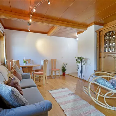 A cozy living room with a gray sofa and a wooden cabinet. The room is bright and friendly, with a table and chairs in the background.