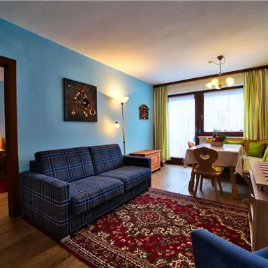 Ein gemütliches Wohnzimmer mit einem blauen Sofa und einem Esstisch. Fenster mit Vorhängen lassen viel Tageslicht herein.