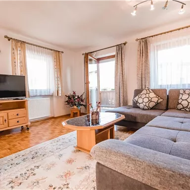 Ein helles Wohnzimmer mit einem grauen Sofa und einem Couchtisch. Auf der anderen Seite steht ein Fernseher auf einem Holzschrank, und große Fenster sorgen für viel Tageslicht.