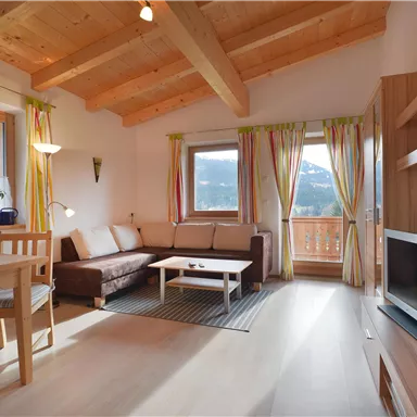 Ein gemütliches Wohnzimmer mit Holzbalkendecke und großen Fenstern. Das Zimmer ist hell und freundlich eingerichtet, mit einem Sofa, Tisch und Pflanzen.