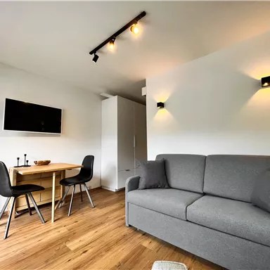 Ein modernes Wohnzimmer mit einem grauen Sofa und einem Holztisch. An der Wand hängt ein Fernseher und der Boden ist aus Holz.