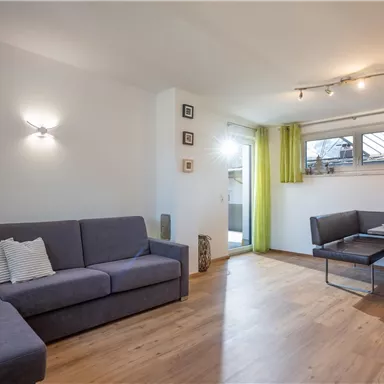 A modern living room with a gray couch and a table for four people. Large windows with green curtains let in plenty of light.