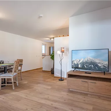 A modern living room with wooden flooring and cozy furnishings. A television is placed on a wooden cabinet, while a dining table with chairs is visible in the background.