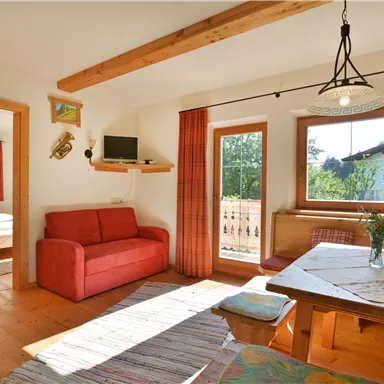 Ein gemütliches Wohnzimmer mit rotem Sofa und Esstisch. Große Fenster bieten Aussicht auf die Natur und lassen viel Licht herein.