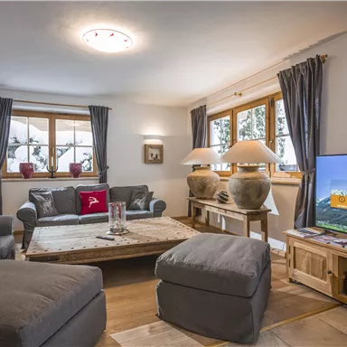 Ein gemütliches Wohnzimmer mit komfortablen Sofas und einem großen Fernseher. Große Fenster lassen viel Licht hinein und schaffen eine einladende Atmosphäre.