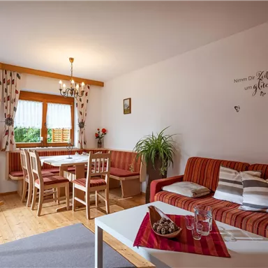 Ein gemütliches Wohnzimmer mit einem roten Sofa und einem Essbereich. Große Fenster lassen viel Licht herein und eine Pflanze sorgt für eine frische Atmosphäre.