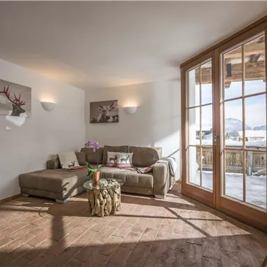 A cozy living room with a gray sofa and large windows leading to a balcony. The room features hardwood flooring and modern decoration.