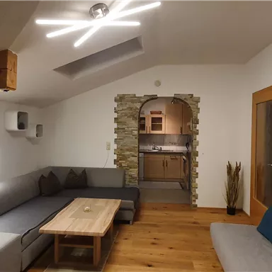 A modern living room with gray sofas and a wooden table. In the background, there is an open kitchen and a piece of art featuring a forest landscape on the wall.