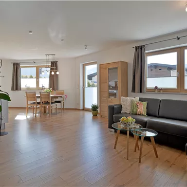 Ein modernes Wohnzimmer mit einem schwarzen Sofa und Holzfußboden. Große Fenster lassen viel Licht herein und es gibt einen Essbereich im Hintergrund.