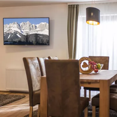Ein einladender Essbereich mit einem Holztisch und gemütlichen Stühlen. An der Wand hängt ein Fernseher mit einem Bild von schneebedeckten Bergen.