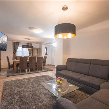 Ein modernes Wohnzimmer mit einem grauen Sofa und einem gläsernen Tisch. Im Hintergrund ist ein Esstisch mit Stühlen und ein Fernseher zu sehen.