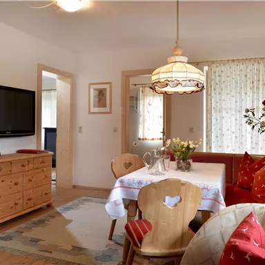 A cozy living space with a dining table and colorful cushions on the couch. A TV hangs on the wall and large windows let in plenty of light.