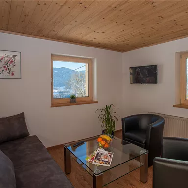A cozy living room with a gray couch, two black chairs, and a glass table. Large windows provide a view of the mountains and plenty of natural light.